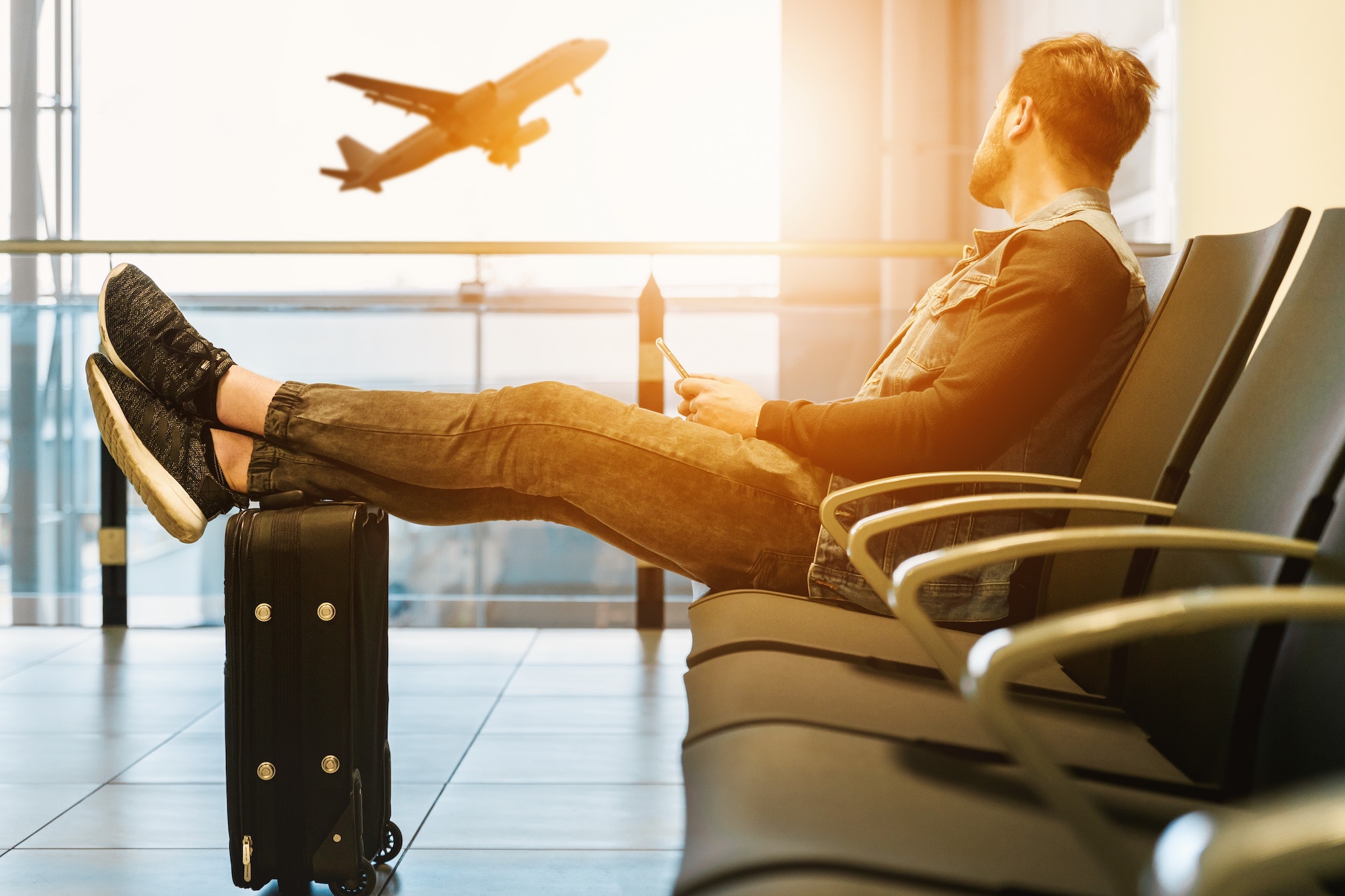 man sitting on gang chair with feet on luggage looking at airplane ready to go with travel health